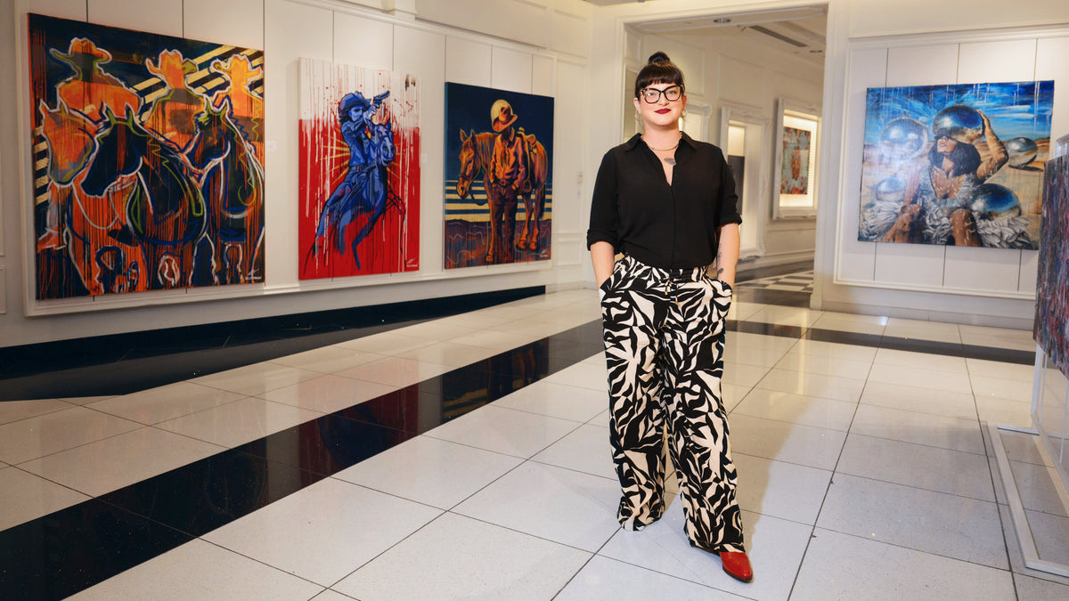 Kelly O'Brien standing in a white gallery hall beside three of her large Western paintings